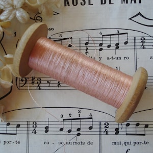Pode incluir: Uma bobina de madeira com fio rosa pálido repousa sobre um fundo de partituras antigas. As extremidades de madeira da bobina emolduram o fio, que está bem enrolado. Delicados detalhes florais são visíveis perto da bobina, adicionando um toque de suavidade.