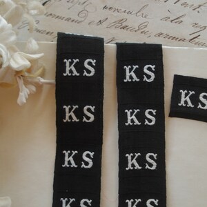 May include: Six black fabric labels with white embroidered letters spelling "KS" in a serif font. The labels are arranged in two vertical rows of three labels each.