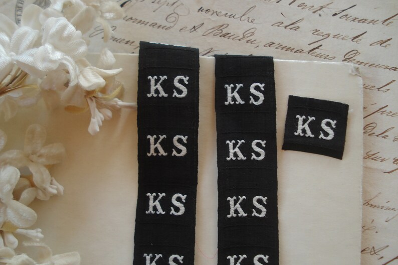 May include: Six black fabric labels with white embroidered letters spelling "KS" in a serif font. The labels are arranged in two vertical rows of three labels each.
