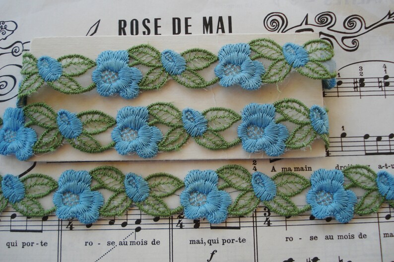 May include: Two rows of blue and green floral lace trim. The lace features a delicate floral design with blue flowers and green leaves.