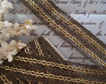 May include: Close-up of gold and brown metallic trim with a delicate, intricate design. The trim is arranged in overlapping layers on a white surface.