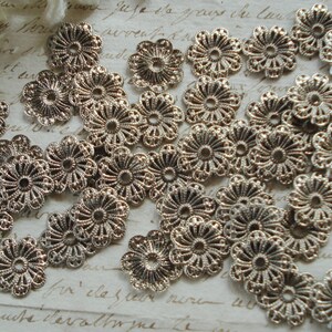 May include: A collection of silver metal flower-shaped embellishments with intricate designs. The embellishments are scattered on a white surface with handwritten text.