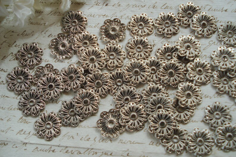 May include: A collection of silver metal flower-shaped embellishments with intricate designs. The embellishments are scattered on a white surface with handwritten text.