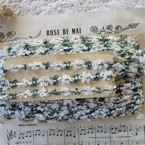 May include: A spool of white and green trim with a label that reads "ROSE DE MAI". The trim is made of a delicate, ruffled fabric and has a vintage look.