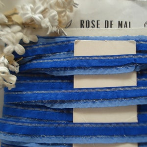 May include: Blue and silver striped ribbon with a frayed edge. The ribbon is folded in layers and has a white label with the text "ROSE DE MAI" on it.