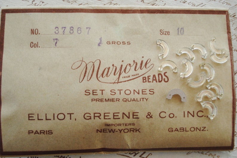May include: A vintage packaging card for Marjorie Beads Set Stones, Premier Quality. The card features the company name, address, and a trademarked logo. The card also includes a size 10 label and a number 37867 label. The card is decorated with a variety of clear glass beads.