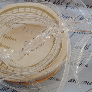 以下が含まれることがあります： 金色のアクセントが付いた白い透き通ったリボンの糸巻き。リボンは、「Farbe:」と「Meter」が印刷された厚紙の糸巻きに巻かれています。糸巻きには「7201」のラベルが貼られています。