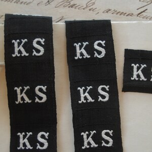 May include: Four black fabric labels with white embroidered letters spelling "KS" on each label. The labels are arranged in two rows of two.