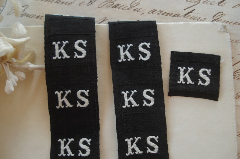 May include: Four black fabric labels with white embroidered letters spelling "KS" on each label. The labels are arranged in two rows of two.