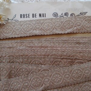 Puede incluir: Primer plano de una cinta decorativa beige con un patr&oacute;n floral repetitivo. La cinta est&aacute; hecha de tejido tejido y tiene una superficie ligeramente texturizada. El texto "ROSE DE MAI" es visible en el fondo.