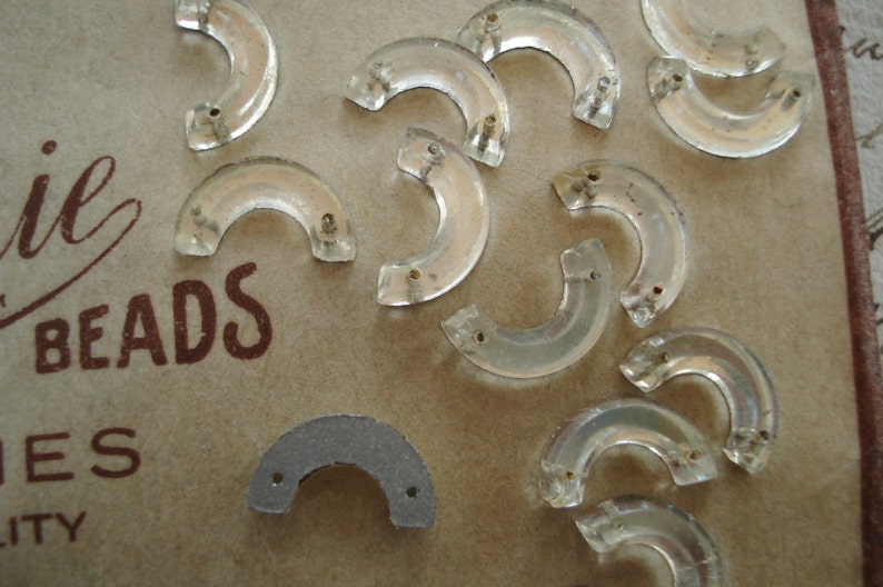 May include: A collection of clear glass beads in the shape of a crescent moon. The beads are arranged on a brown surface with the word "BEADS" printed on it.