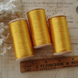 Pode incluir: Três carretéis de fio dourado, cada um enrolado em uma bobina de madeira. Os carretéis estão sobre um pedaço de papel com escrita.