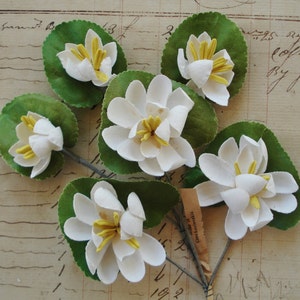 Könnte beinhalten: Sechs künstliche weiße Seerosen mit gelben Zentren und grünen Blättern sind auf einem Vintage-Papierhintergrund angeordnet. Die Blumen bestehen aus Stoff und sind an dünnen Drahtstielen befestigt. Das Papier hat handschriftlichen Text.