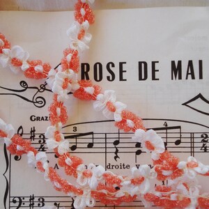 May include: A white and pink crocheted trim with a floral design. The trim is laid out on a sheet of music with the title "ROSE DE MAI".