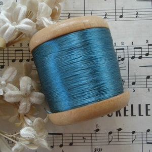 May include: A spool of teal blue thread wound around a wooden bobbin. The spool is sitting on a sheet of music with the word "ORELLE" visible.