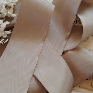 Könnte beinhalten: Ein breites, beiges Satinband mit dezentem Glanz, kunstvoll in fließenden Falten angeordnet. Die Kanten des Bandes sind sauber genäht. Weiße Blumen und Notenblätter bilden einen weichen Hintergrund und erzeugen eine zarte und elegante Ästhetik.