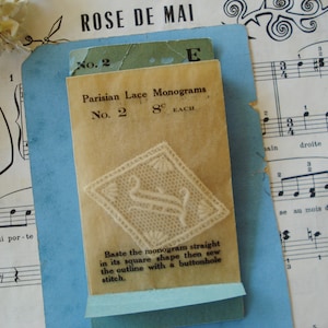 May include: A vintage packet of Parisian Lace Monograms, featuring a delicate white lace design in the shape of the letter "S". The packet is labelled "No. 2" and "8¢ EACH". Instructions for basting and sewing the monogram are included.