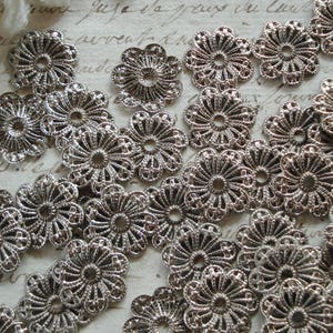 May include: A collection of silver metal filigree embellishments with a floral design. The embellishments are arranged in a random pattern on a white surface.