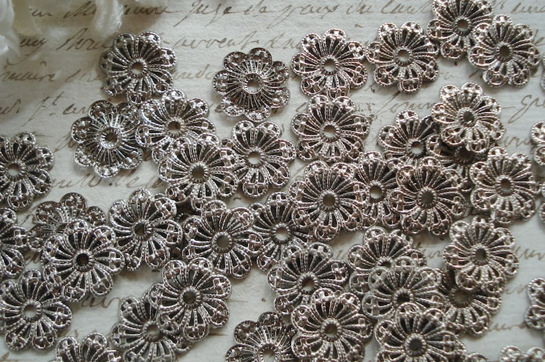 May include: A collection of silver metal filigree embellishments with a floral design. The embellishments are arranged in a random pattern on a white surface.