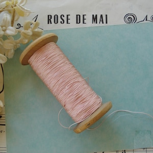 Pode incluir: Uma bobina de fio rosa claro enrolada em torno de uma bobina de madeira. O fio é fino e delicado, e a bobina é de cor marrom claro. A bobina está sobre uma superfície azul claro com as palavras "Rose de Mai" impressas nela.