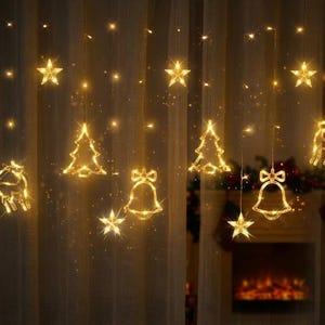 Pode incluir: Uma cortina de luzes LED de cor branca quente com formas decorativas suspensas, incluindo estrelas, árvores de Natal, sinos e uma rena. As luzes estão dispostas em fios verticais, criando uma exibição festiva e brilhante.