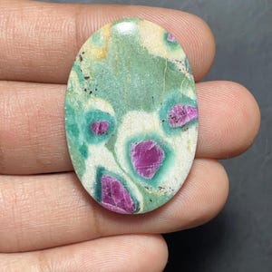 Peut inclure: Une pierre précieuse cabochon de forme ovale présentant une surface marbrée avec un mélange de vert clair, de crème et de blanc. La pierre est incrustée de plusieurs inclusions distinctes rose magenta vif entourées de halos vert foncé.