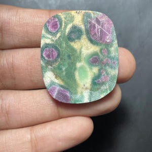 Peut inclure: Une pierre pr&eacute;cieuse plate et polie de forme rectangulaire aux coins arrondis, pr&eacute;sentant un motif tachet&eacute; d'inclusions rose magenta profond incrust&eacute;es dans une matrice de d&eacute;p&ocirc;ts min&eacute;raux vert for&ecirc;t, olive et blanc cass&eacute;.