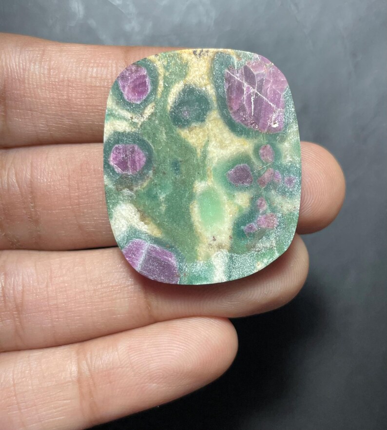 Peut inclure: Une pierre pr&eacute;cieuse plate et polie de forme rectangulaire aux coins arrondis, pr&eacute;sentant un motif tachet&eacute; d'inclusions rose magenta profond incrust&eacute;es dans une matrice de d&eacute;p&ocirc;ts min&eacute;raux vert for&ecirc;t, olive et blanc cass&eacute;.