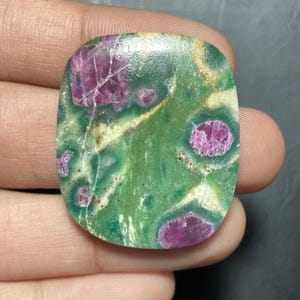 May include: A polished, rectangular-shaped cabochon gemstone featuring a vibrant green base with embedded, irregular magenta-coloured crystal inclusions and thin, light-coloured mineral veins running across the smooth, glossy surface.