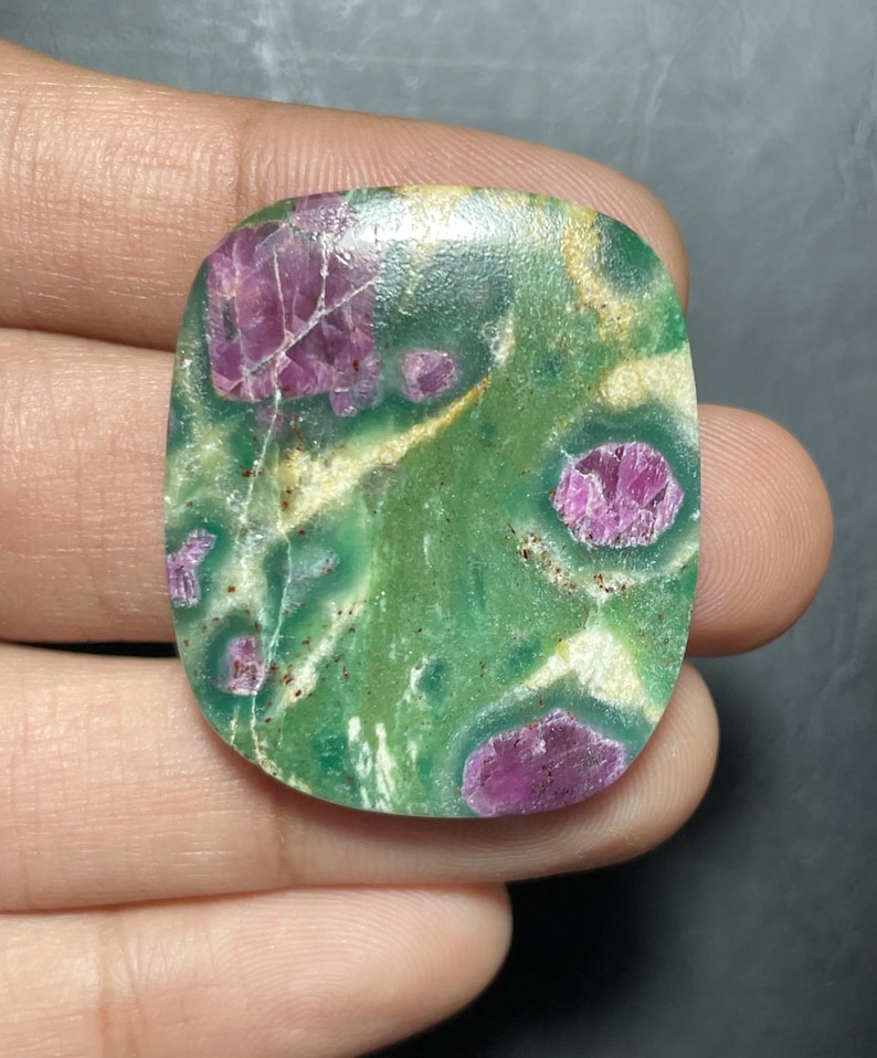 Peut inclure: Une pierre pr&eacute;cieuse cabochon polie de forme rectangulaire pr&eacute;sentant une base vert vif avec des inclusions de cristaux magenta irr&eacute;guliers et de fines veines min&eacute;rales claires traversant la surface lisse et brillante.