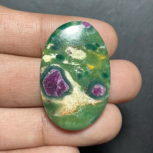 Op de afbeelding: Een ovaalvormige cabochon edelsteen met een glad, gepolijst oppervlak, een gevlekte groene basiskleur, afgewisseld met crèmewitte en gele vlekken, en drie duidelijke, ingebedde diep magenta-kleurige robijninsluitingen.