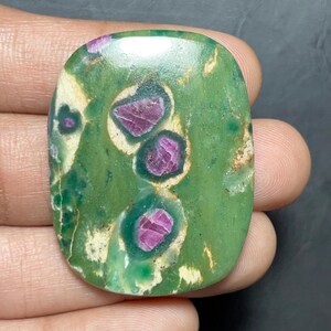 Puede incluir: Una piedra preciosa cabujón ovalada y pulida que presenta una matriz de zoisita de color verde vibrante con inclusiones de rubí magenta intenso. La piedra muestra variaciones de color naturales, incluyendo manchas blanco crema y vetas minerales verde oscuro.