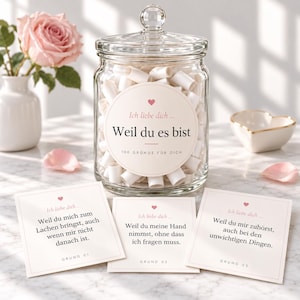 Könnte beinhalten: Ein durchsichtiges Glas mit Deckel, gefüllt mit kleinen weißen Papierrollen. Das Etikett trägt die Aufschrift: Ich liebe dich ... Weil du es bist 100 GRÜNDE FÜR DICH. Drei quadratische Karten davor zeigen deutschen Text.