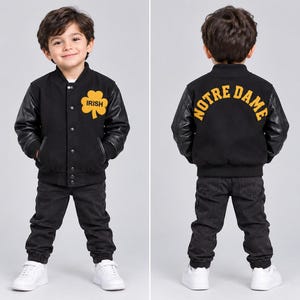 May include: A black varsity jacket with black sleeves, a front yellow shamrock graphic with the text IRISH, and a back yellow arched text reading NOTRE DAME. The jacket features a snap-button closure, ribbed collar, cuffs, and hem.