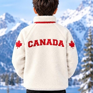 Peut inclure: Une veste en polaire crème à manches longues avec un col montant et un liseré rouge. Le dos porte l'inscription "CANADA" en lettres rouges audacieuses, flanquée d'une petite icône de feuille d'érable rouge sur chaque manche.
