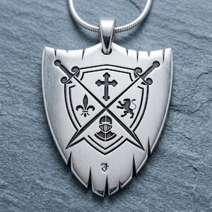 May include: A silver-toned pendant shaped like a shield with a jagged bottom edge, suspended from a silver chain. The surface features an engraved design of two crossed swords, a cross, a fleur-de-lis, a lion, and a knight's helmet.