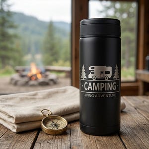 Könnte beinhalten: Ein mattschwarzer, zylindrischer Isolierbecher mit einer Lasergravur eines Wohnmobils zwischen zwei Tannen sowie dem Schriftzug "CAMPING" und "LIVING ADVENTURE". Ein goldfarbener, runder Kompass liegt auf einem gefalteten beigen Tuch.