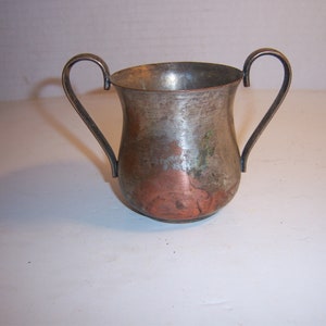 May include: A small, aged metal cup with two curved handles. The cup has a hammered texture with a mix of silver, copper, and green hues. The cup's shape is wider at the top and tapers towards the base.