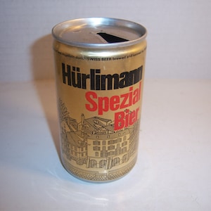 May include: A vintage Hürlimann Spezial Bier can. The can is gold with black and red text, and a detailed illustration of a European town. The top of the can is silver, with a pull-tab opening.
