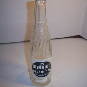 May include: A clear glass bottle with a textured surface. The bottle has a black and white label that reads "Mission Beverages Naturally Good".