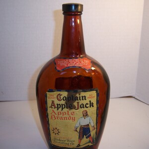 May include: A brown glass bottle with a label that reads "Captain Apple Jack Apple Brandy 90 Proof" and features a cartoon pirate holding a sword. The bottle is one quart and is made by Hickory Town Distilling Co.