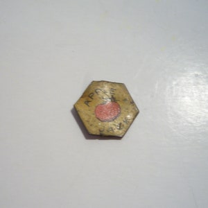 May include: A small, hexagonal tag with the word "APPLE" and a red apple illustration. The tag is yellowed with age and has the word "JAM" below the apple. The background is a plain white surface.