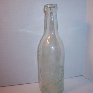 May include: A clear glass bottle with a textured surface. The bottle has a rounded bottom and a narrow neck. The bottle is empty and appears to be vintage.