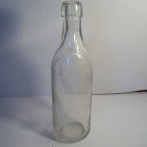 May include: A clear glass bottle with a rounded bottom and a narrow neck. The bottle has a raised circular design around the middle with text that is difficult to read.