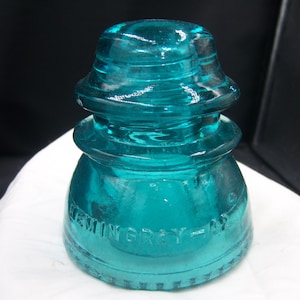 May include: A teal-colored glass insulator with a rounded top and base. The insulator has a ribbed design and the text "HEMINGRAY - NY" is embossed on the side. The object is sitting on a white surface.