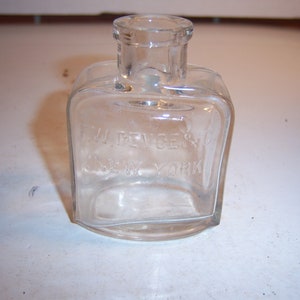 May include: A clear glass ink bottle with a square body and a round neck. The bottle has the text "F.W. DEVOE & C.O. NEW YORK" etched into the side.