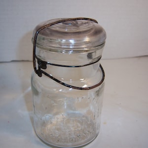 May include: A clear glass jar with a glass lid and a wire handle. The jar has embossed text and a metal wire closure. The lid is secured by a wire bail. The jar is likely for preserving food.