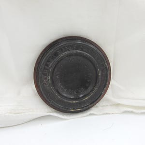 May include: A round, dark brown object with a raised rim and embossed text. The object appears to be a lid or stopper, possibly for a bottle or container. The text is difficult to read, but the object has a vintage or antique appearance.