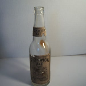 May include: A clear glass bottle with a label that reads "Pickwick Ale". The label is faded and has some brown staining. The bottle is empty.