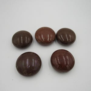 May include: Five smooth, round, dark brown stones with a glossy finish are arranged on a white surface. The stones vary slightly in color, with some appearing more reddish-brown.